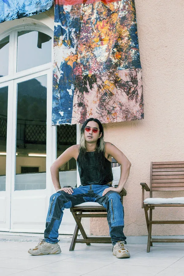 Korakrit Arunanondchai seated on a terrace beneath a painted denim work